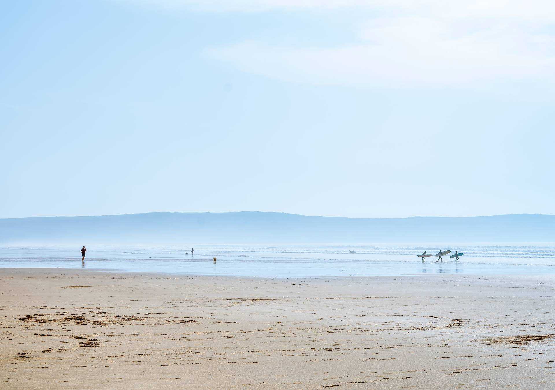 A large stretch of sandy beach with people visible in the distance. One is walking a dog, three people are walking into the sea with surfboards.
