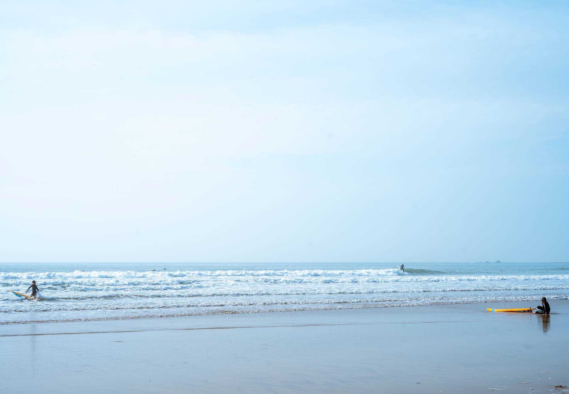A surfer heads out into the sea. Another rests on the beach with their surfboard in front of them.