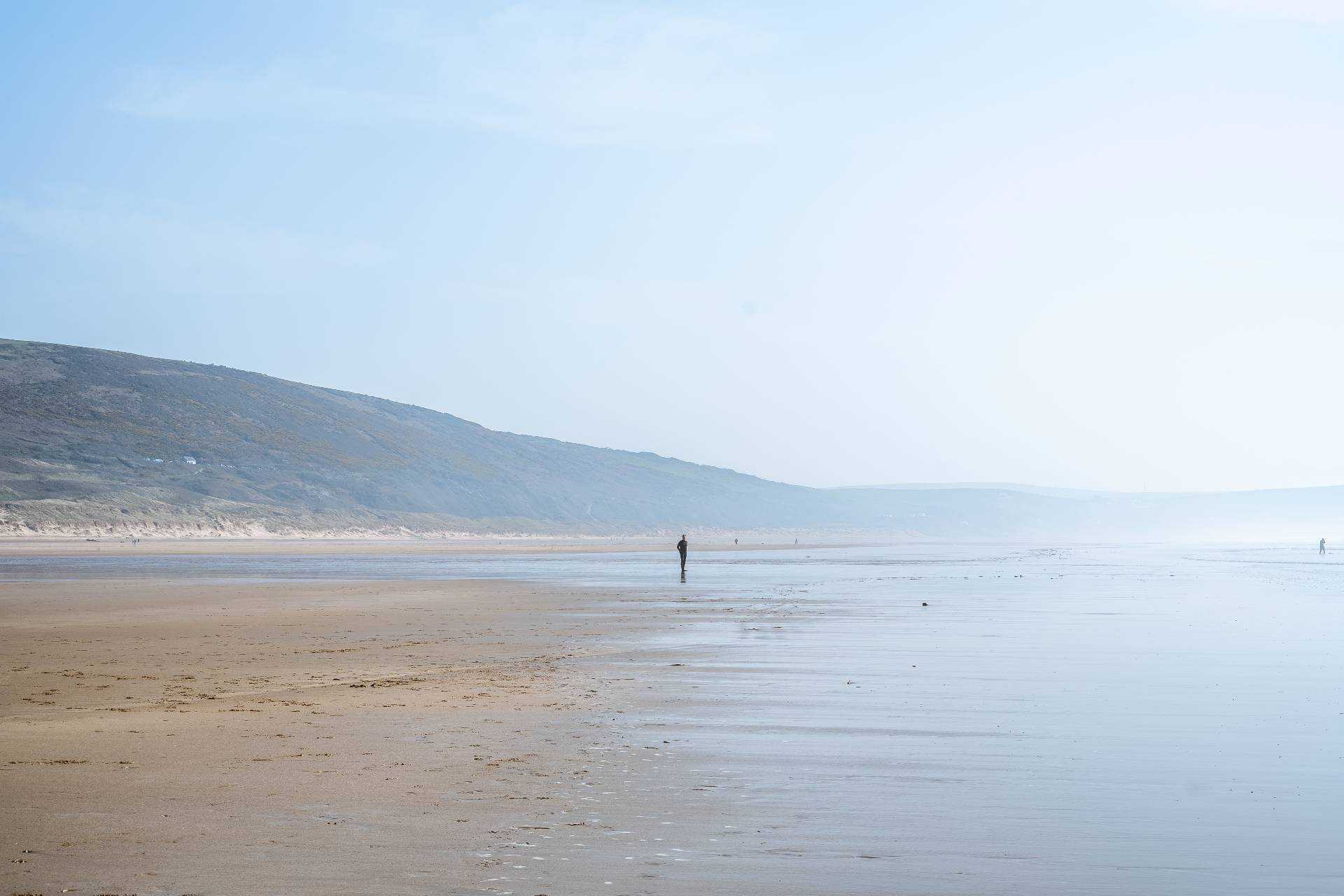 A single person standing on a beach with clifftops in the background.