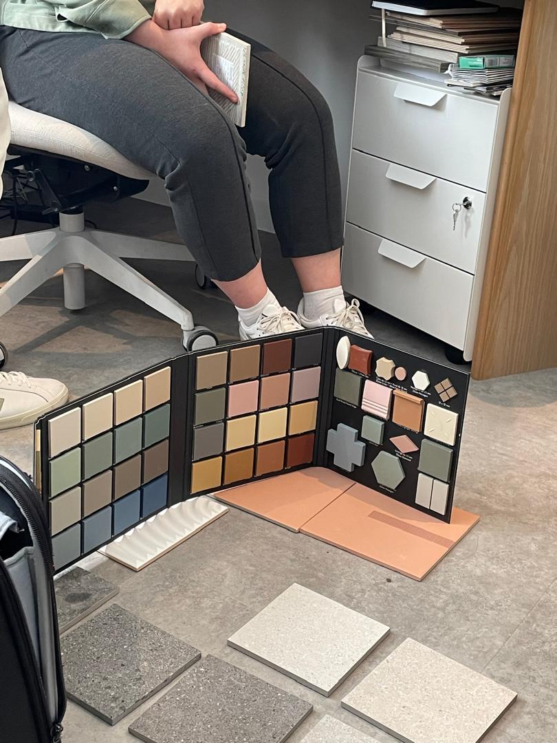 Person seated at desk with open colour sample folders displaying various paint or material swatches on floor in office setting.