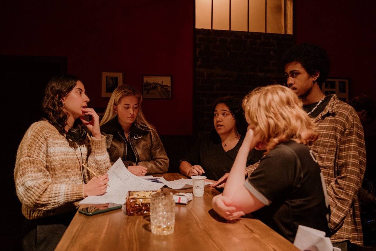 Five people seated around wooden table in dimly lit room, papers scattered, warm lighting from overhead fixture.
