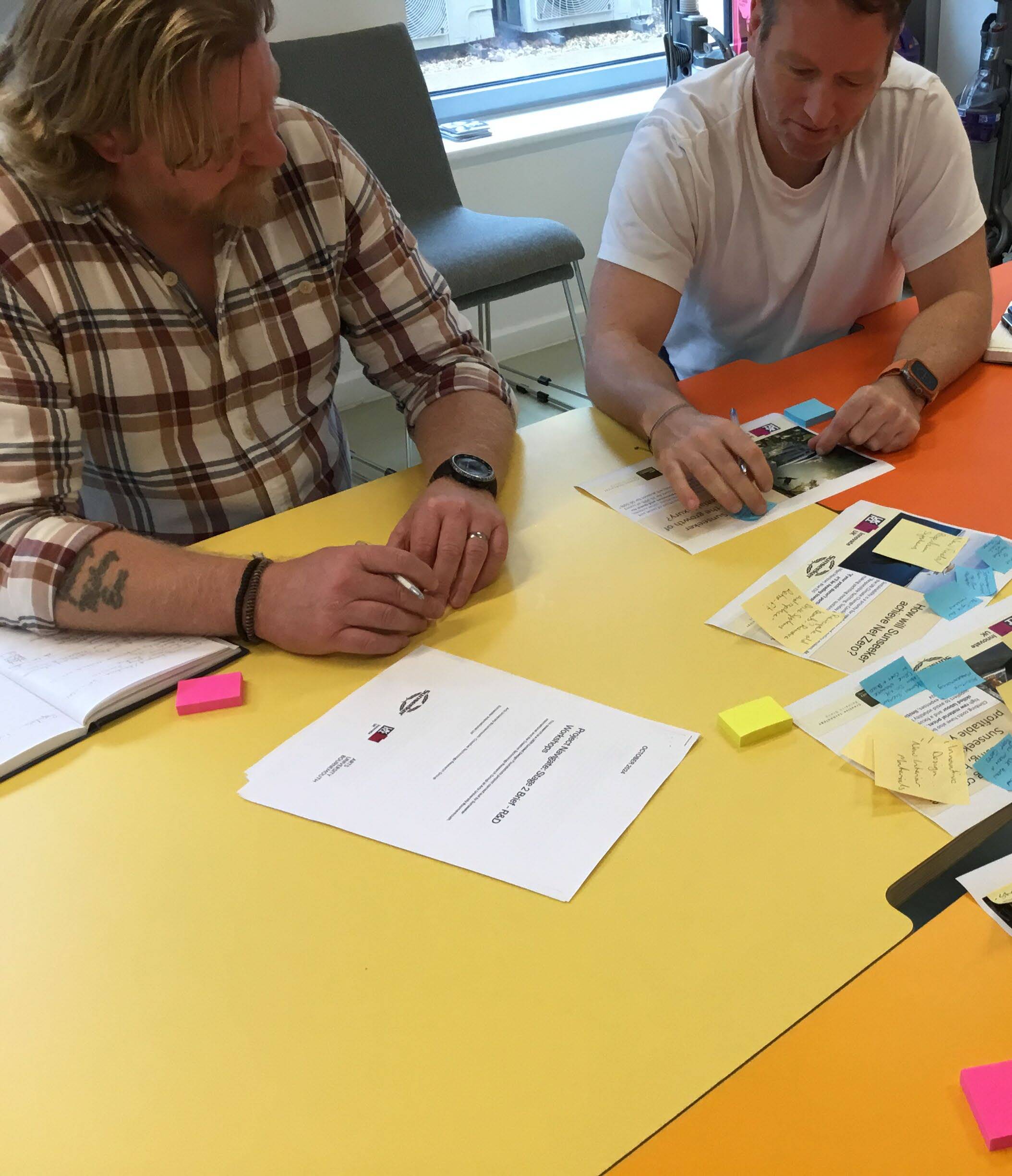 Two people at yellow table with papers, sticky notes, and materials. Woman in checked shirt, man in white t-shirt collaborating on documents.