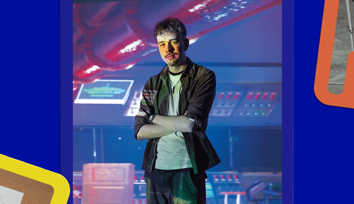 Man with crossed arms wearing dark jacket and light shirt stands against blue wall with colourful projected graphics behind him.