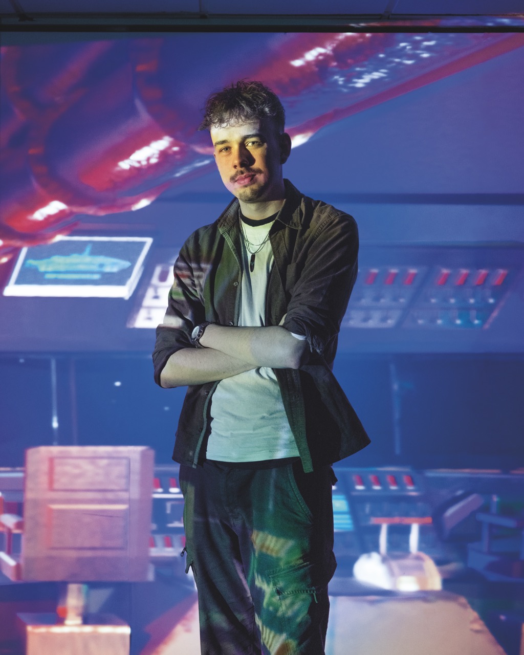 Student stood with crossed arms and casual clothing, colourful projection beaming onto them and the background in blue and yellow resembling a control panel.