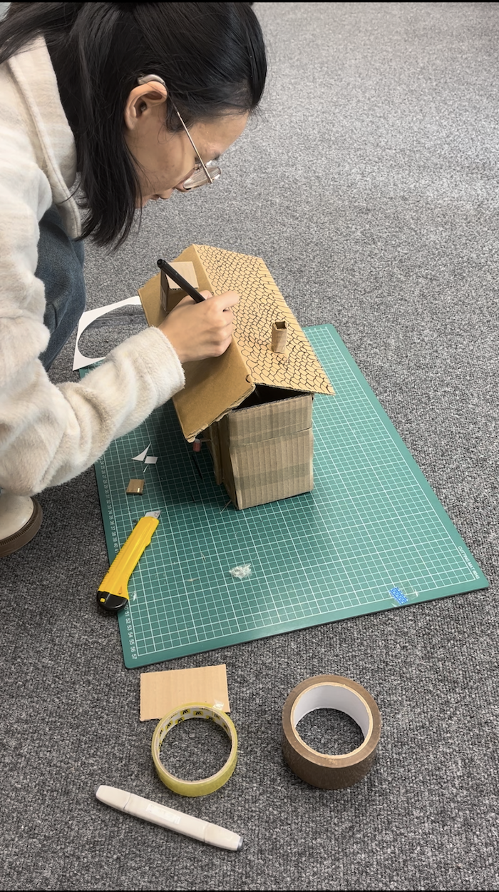 An MA Fine Art student illustrating roof tiles onto a cardboard model of a house, resting on a green cutting board with art supplies surrounding.