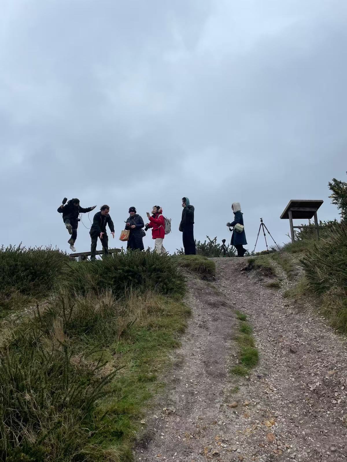 A group of six MA Fine Art students walking along the top of a grassy hill with camera equipment around them and a cloudy grey sky behind them.