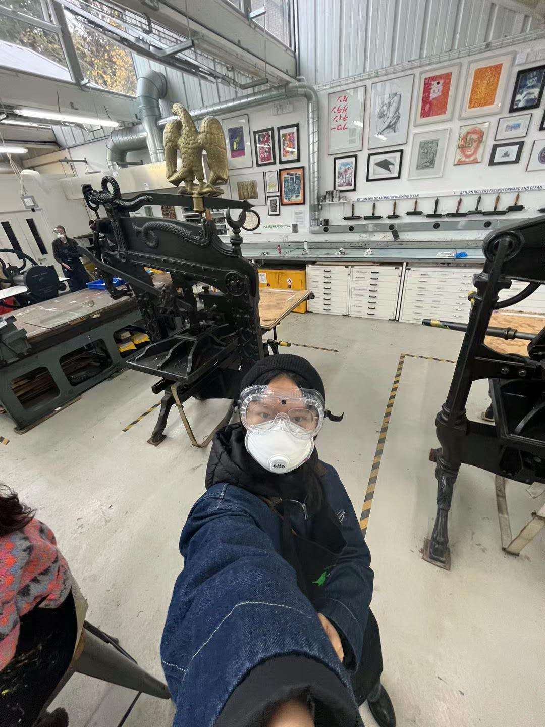 MA Fine Art student X Chen wearing an protective eye and face mask taking a selfie with two printing presses in the background, in the print room at Arts University Bournemouth.