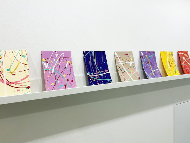 Row of colourful abstract paintings on white shelf against white wall. Works feature paint splatters in pink, blue, beige, purple, yellow, and red.