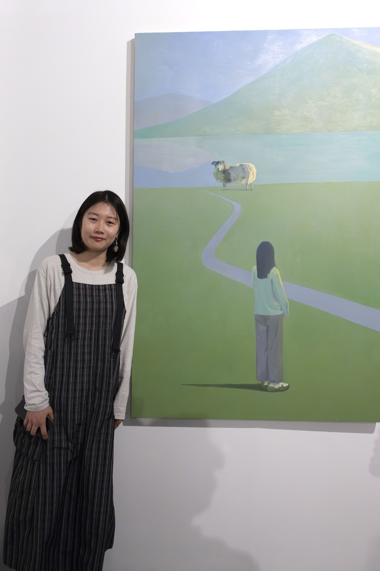 Artist X Wand standing next to a canvas of their painting showing a person with their back to the viewer, looking at a sheep at the end of a path, with a mountain in the distance.