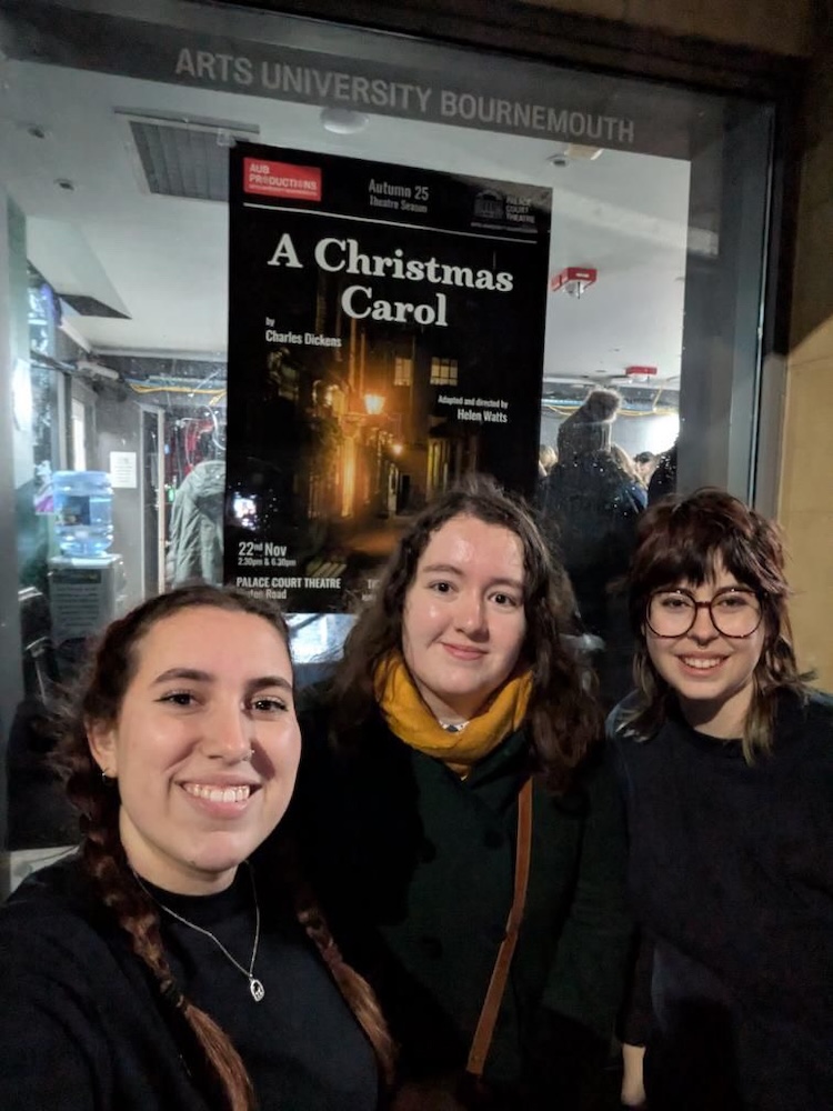 Three people pose in front of a poster for "A Christmas Carol" by Charles Dickens displayed on a glass window. The poster includes details about the show at Arts University Bournemouth, and the background shows a dimly lit venue with a hint of merchandise.