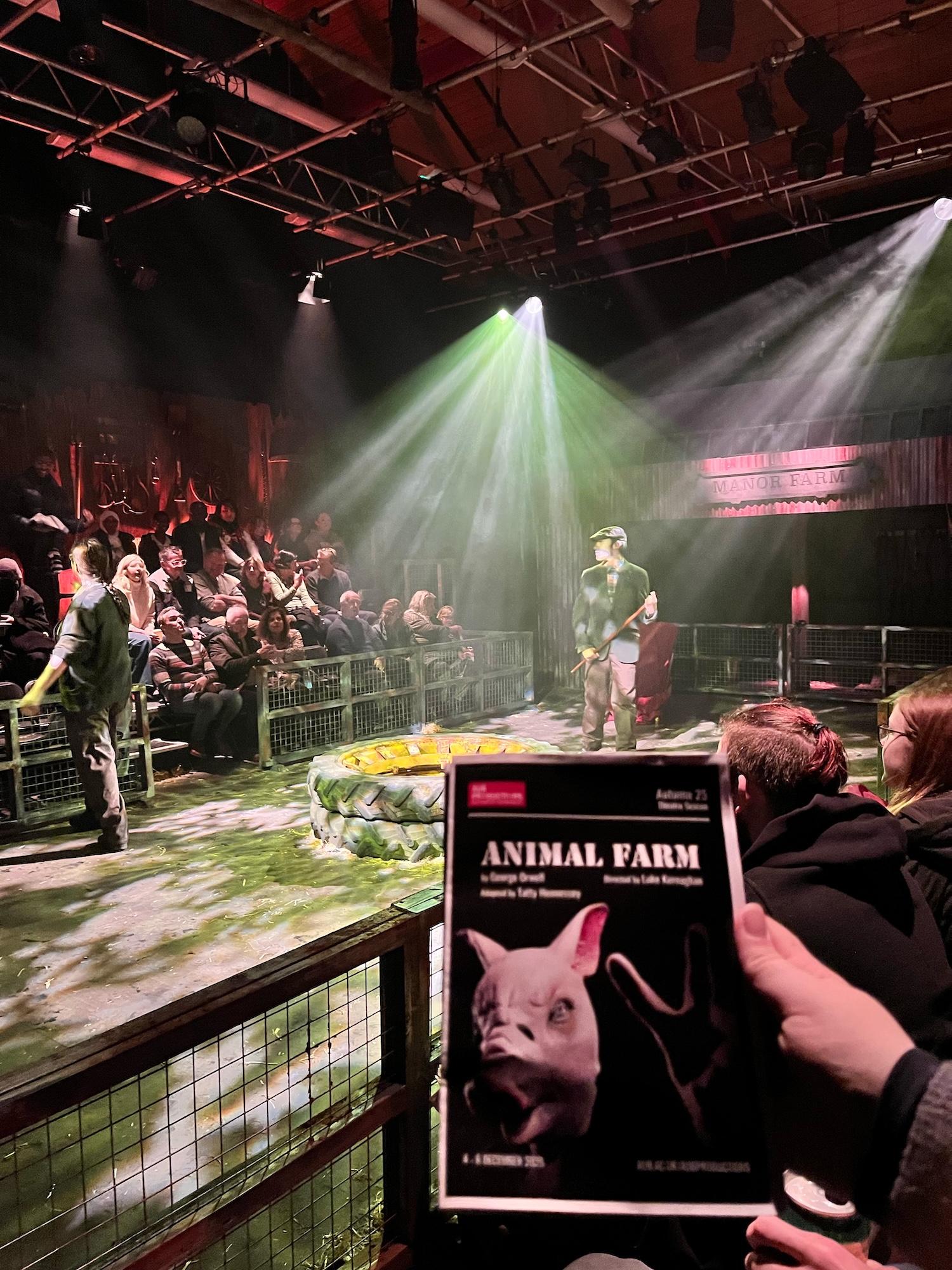 A stage set for a performance of 'Animal Farm' features a man in a hat and grey coat, standing near a circular platform in a dimly lit venue. The audience is seated in the background, illuminated by beams of light.