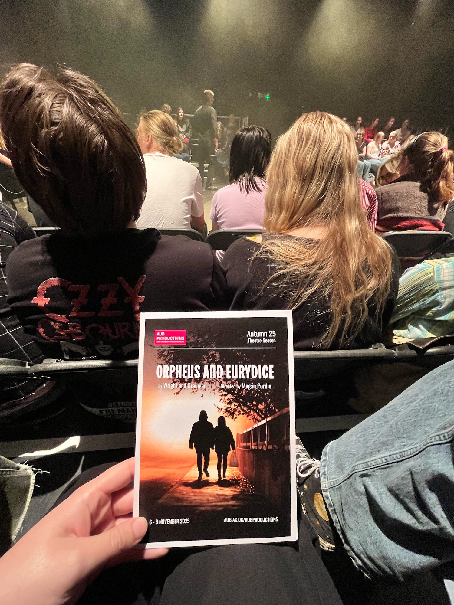 A person holding a programme for 'Orpheus and Eurydice' in a dimly lit theatre. In the background, audience members are seated, some with long hair, while a performer is visible on stage.