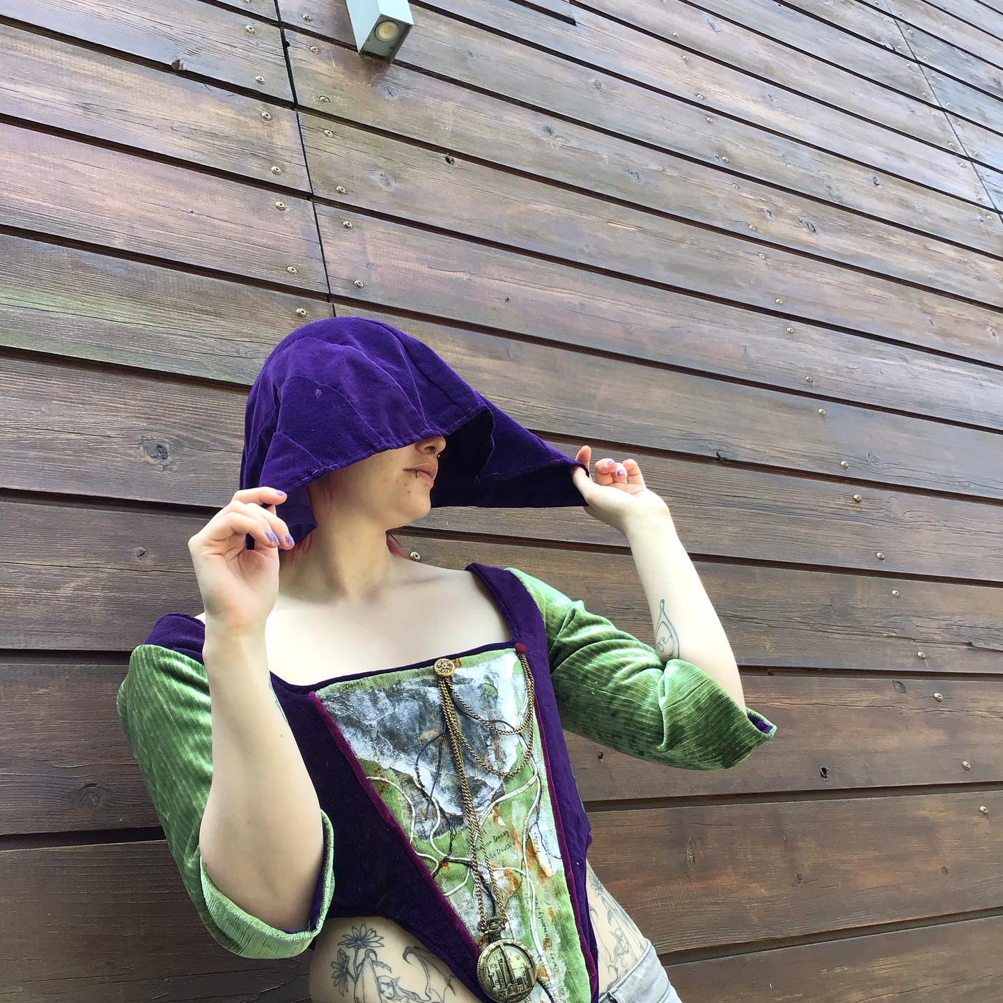 A person in a green and purple corset lifts a purple hood over their face, standing before a wooden plank wall.