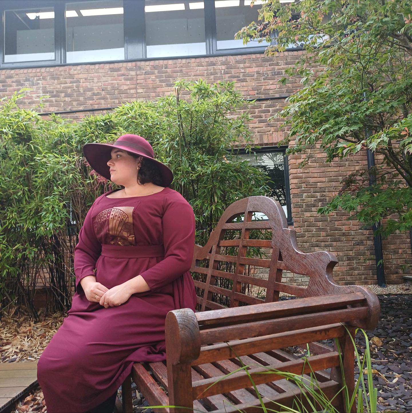 A woman in a burgundy dress and wide-brimmed hat sits on a wooden bench in a landscaped courtyard beside bamboo and a brick building.