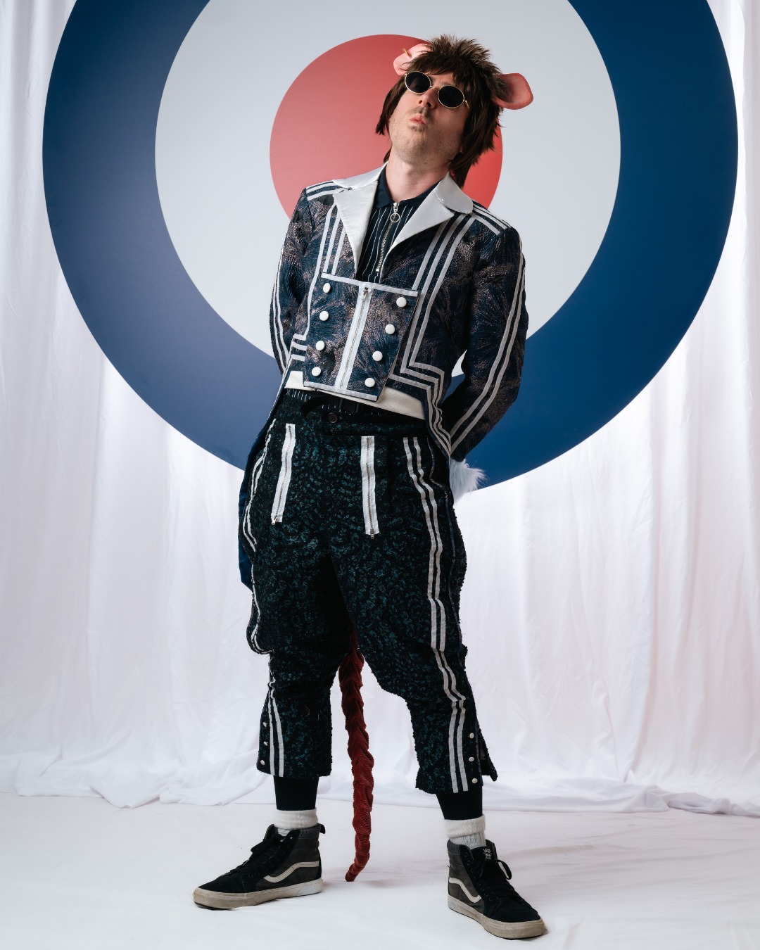 A person in a patterned outfit, sunglasses and dog ears poses in front of a large target-style backdrop with white drapes.