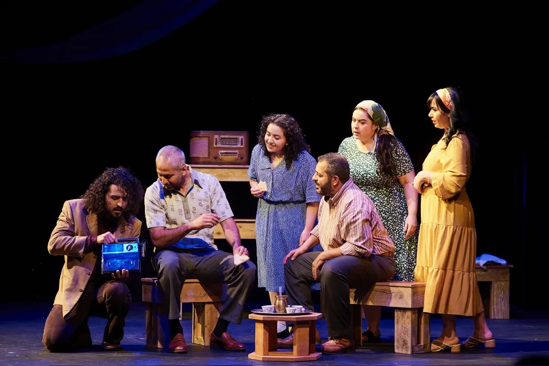Five people on a stage, with two seated on benches while two women and one man stand behind them, and a small table with cups in front.
