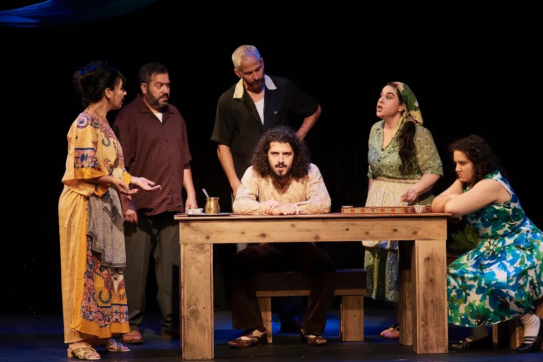 Five people stand and sit around a wooden table on a dark stage, with one man seated at the centre and others gathered beside him.