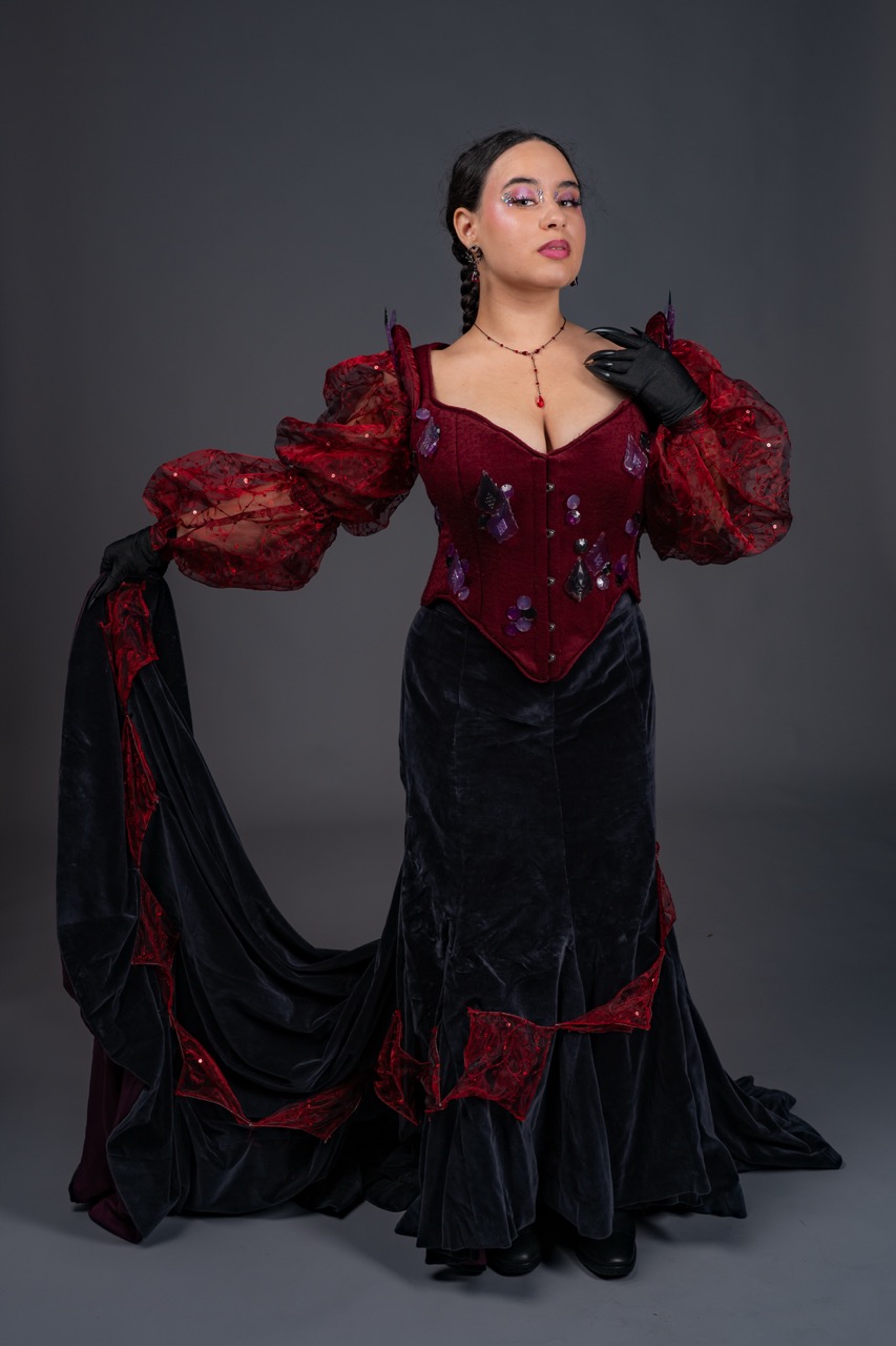 A woman in a red corset-style dress with sheer puff sleeves and a long black velvet skirt poses against a grey studio backdrop, holding up the trailing fabric.