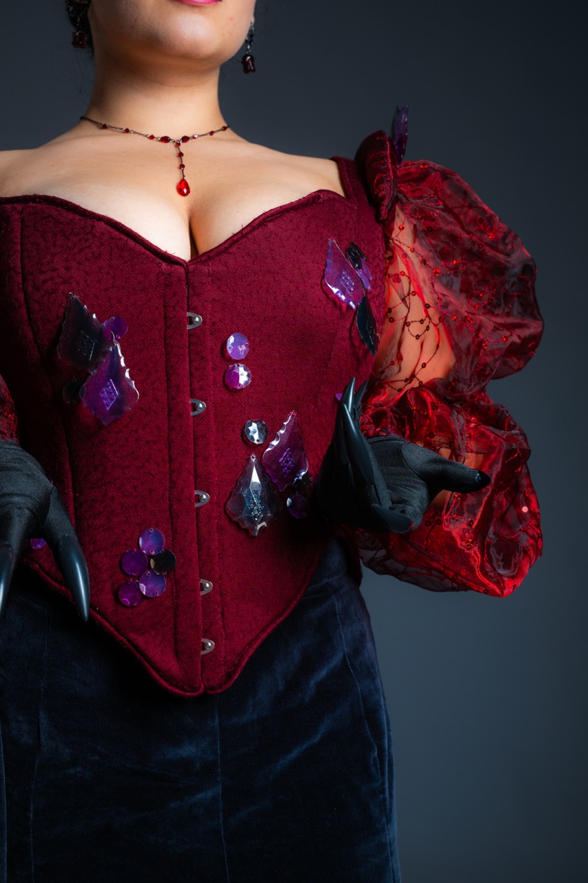 A woman wearing a burgundy corset top with red lace puff sleeves and black gloves, against a dark plain background.