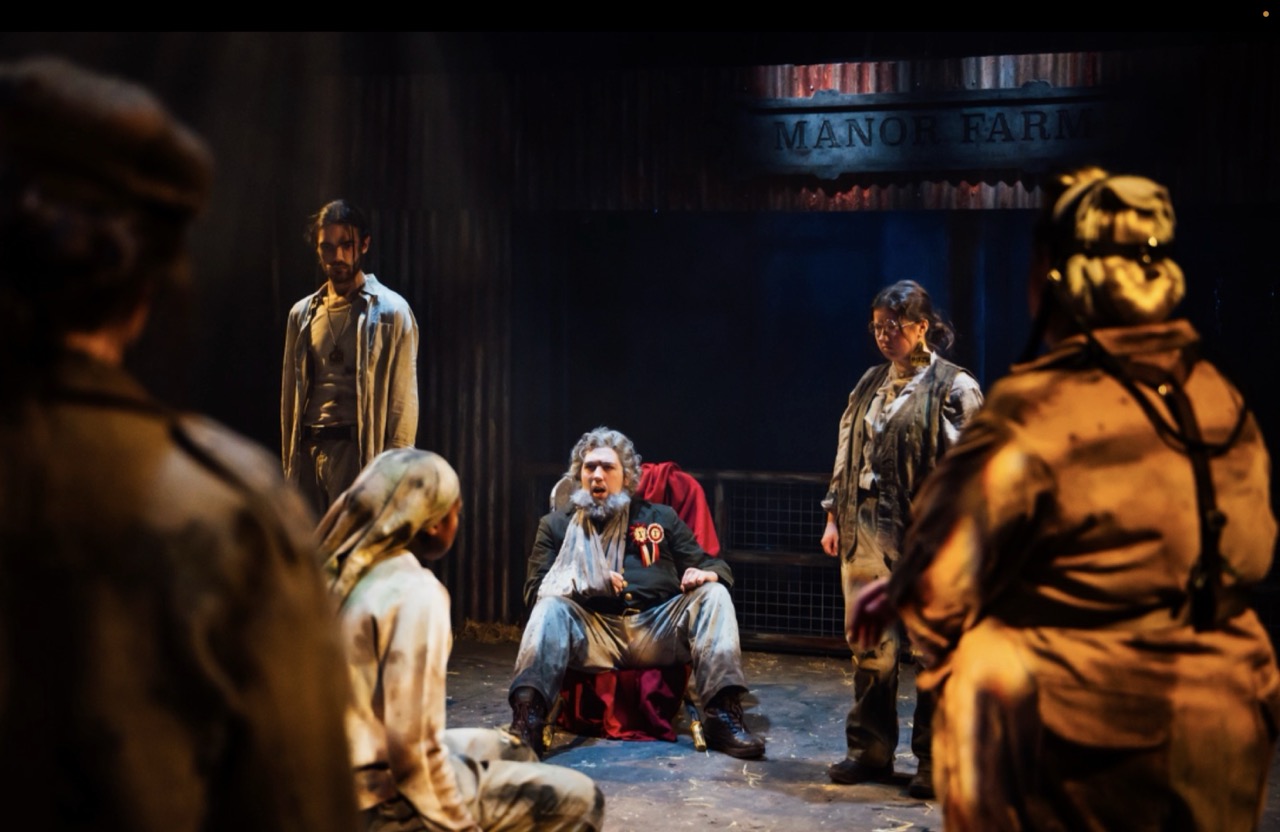 A bearded man sits on a red-covered chair at the centre of a dimly lit stage, surrounded by four other figures in work clothes, with curtains and a sign reading 'Manor Farm' in the background.