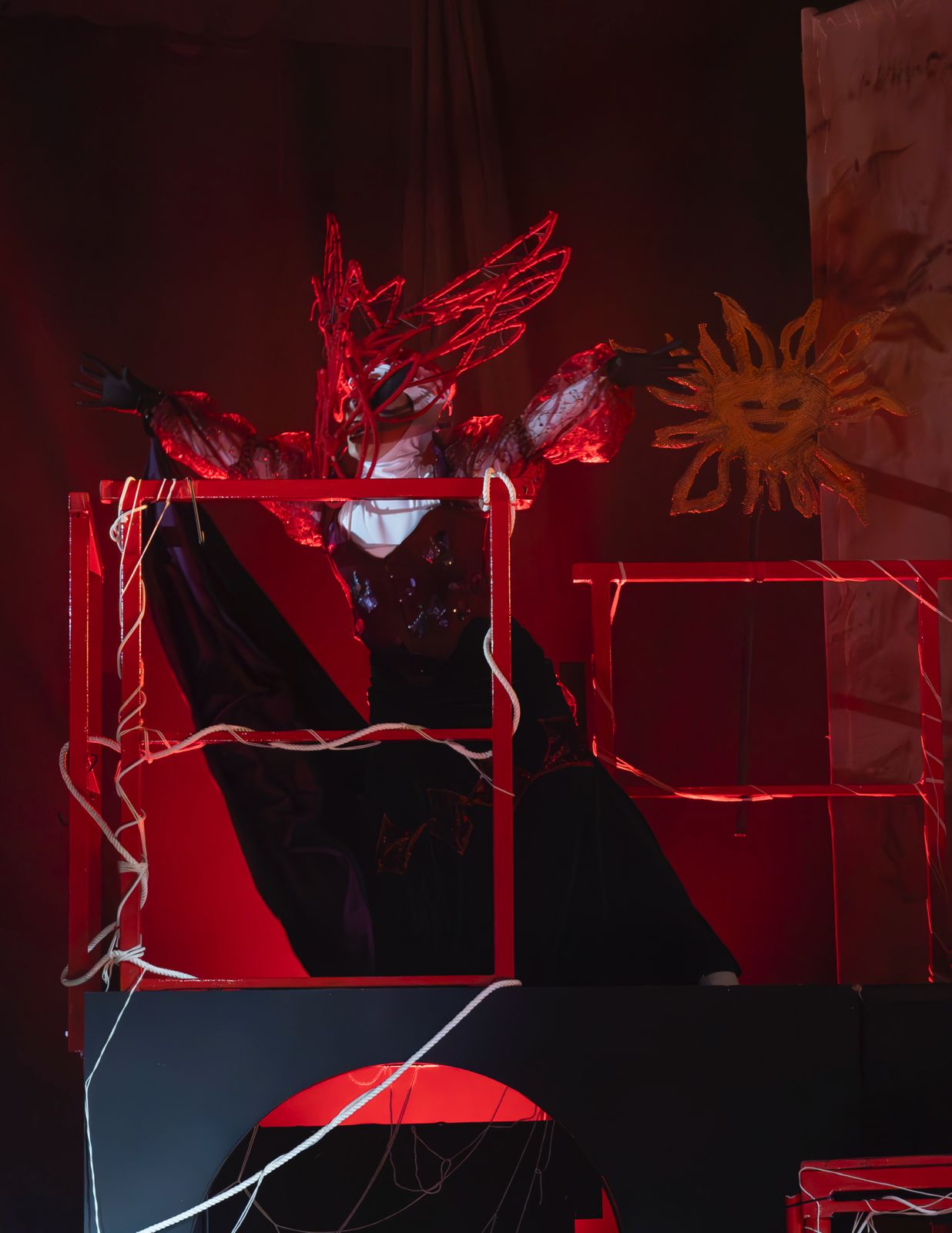 A performer in a red spiked costume stands behind red metal frames, with a painted sun on the backdrop and black drapery behind.