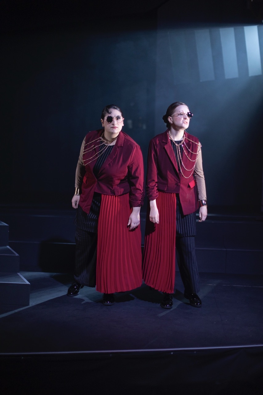 Two performers in matching red velvet jackets and pleated skirts with gold chain necklaces on dark stage with blue lighting.