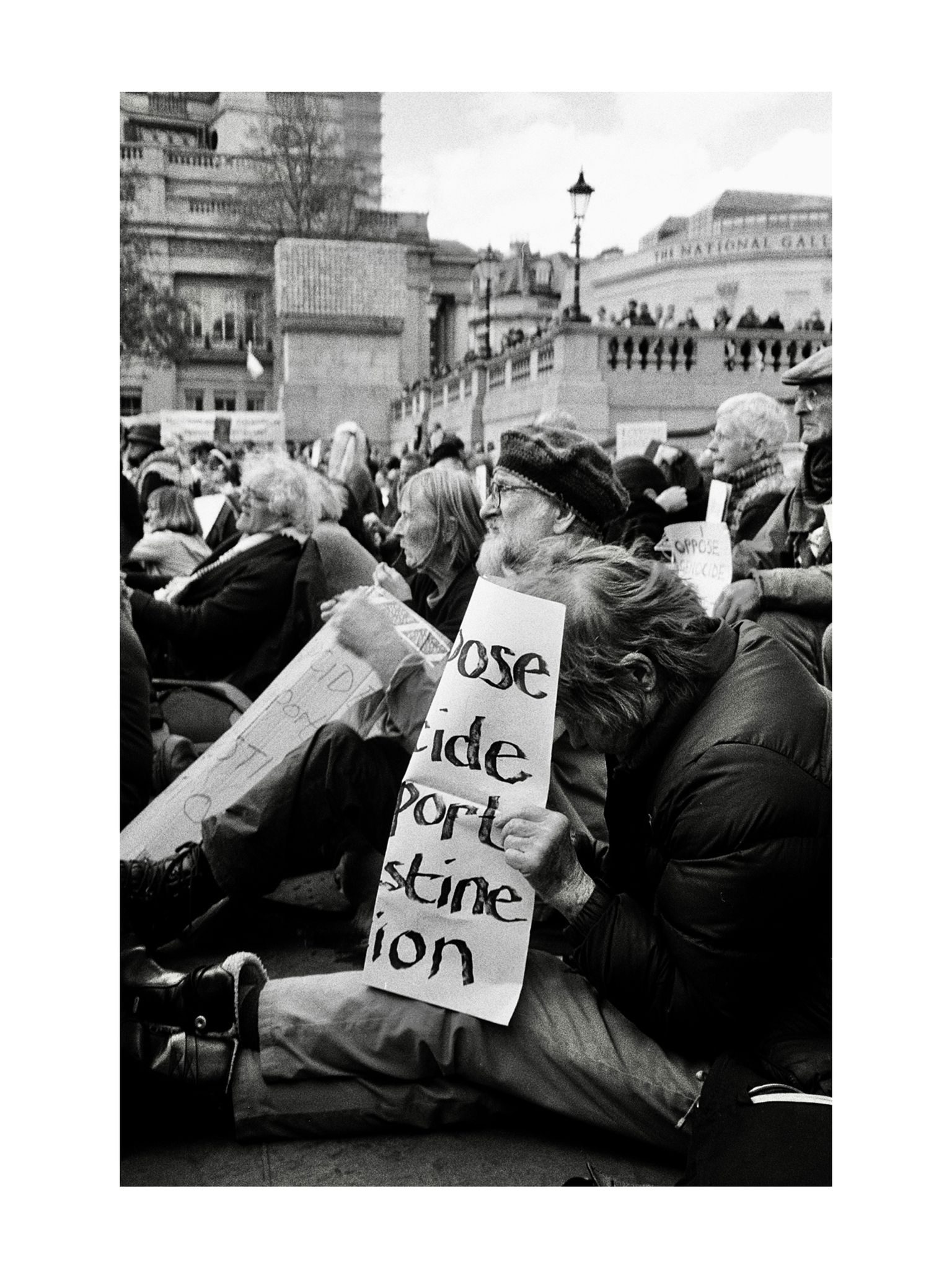 Black and white photograph of Palestine Liberation protestors sitting on a London bridge. One protestor hides their head behind their sign.