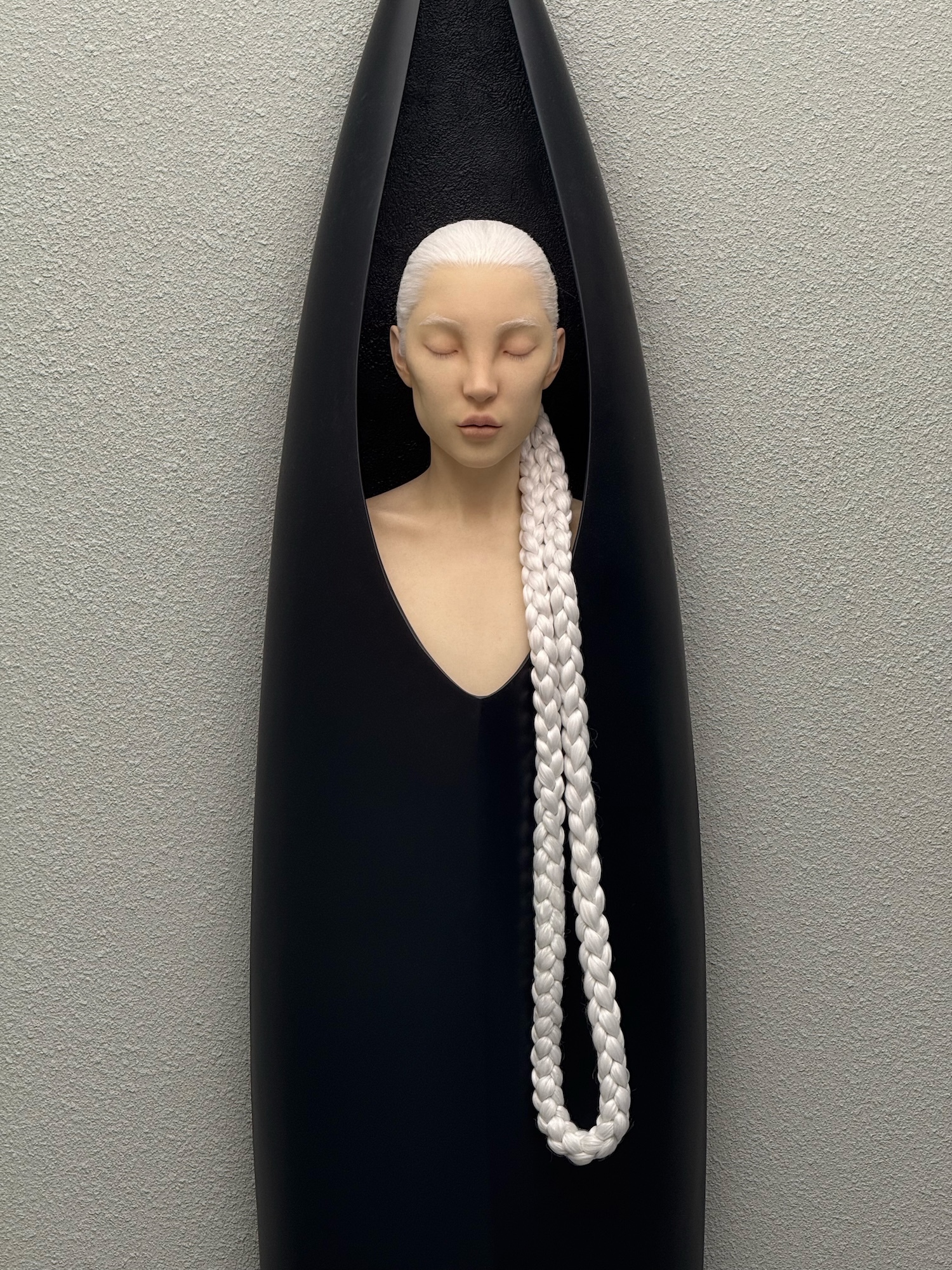 Pale mannequin head with closed eyes in black triangular frame against grey textured wall, wearing long white beaded necklace.
