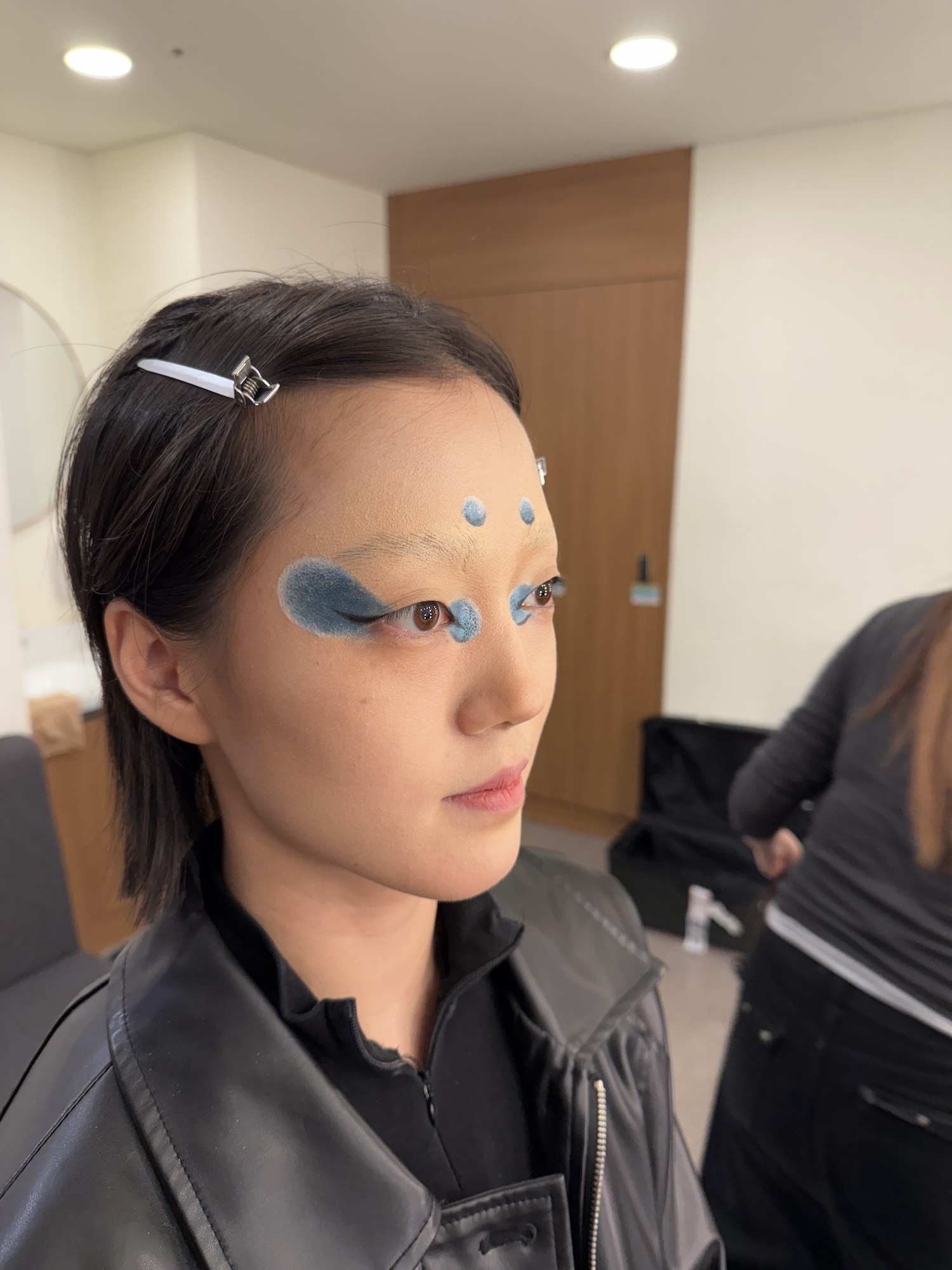 Woman with blue face paint markings around eyes wearing black leather jacket in modern interior with white walls and wood panelling.