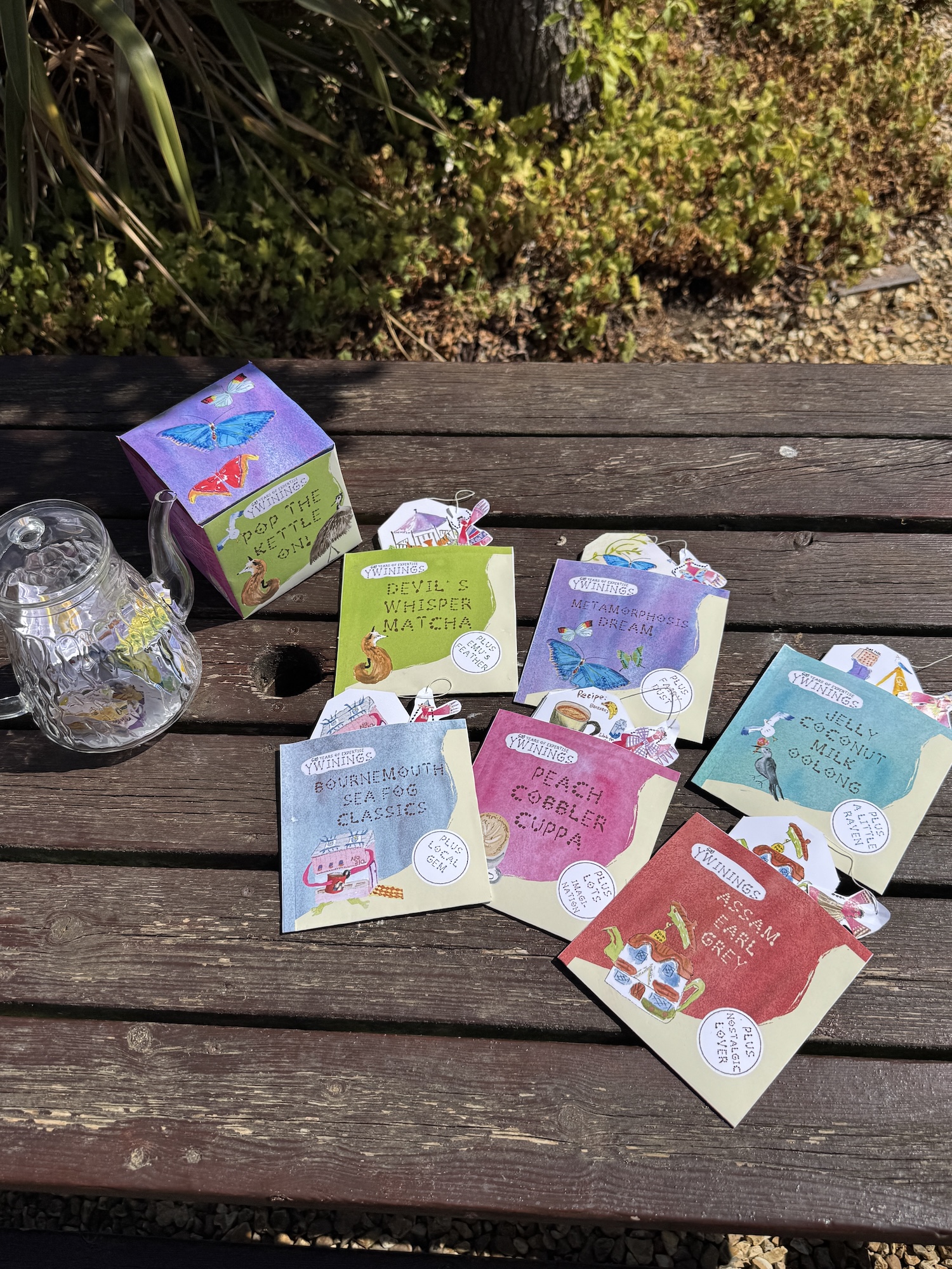 A glass jar and several colourful tea sachet boxes are arranged on a wooden picnic table, with greenery in the background.