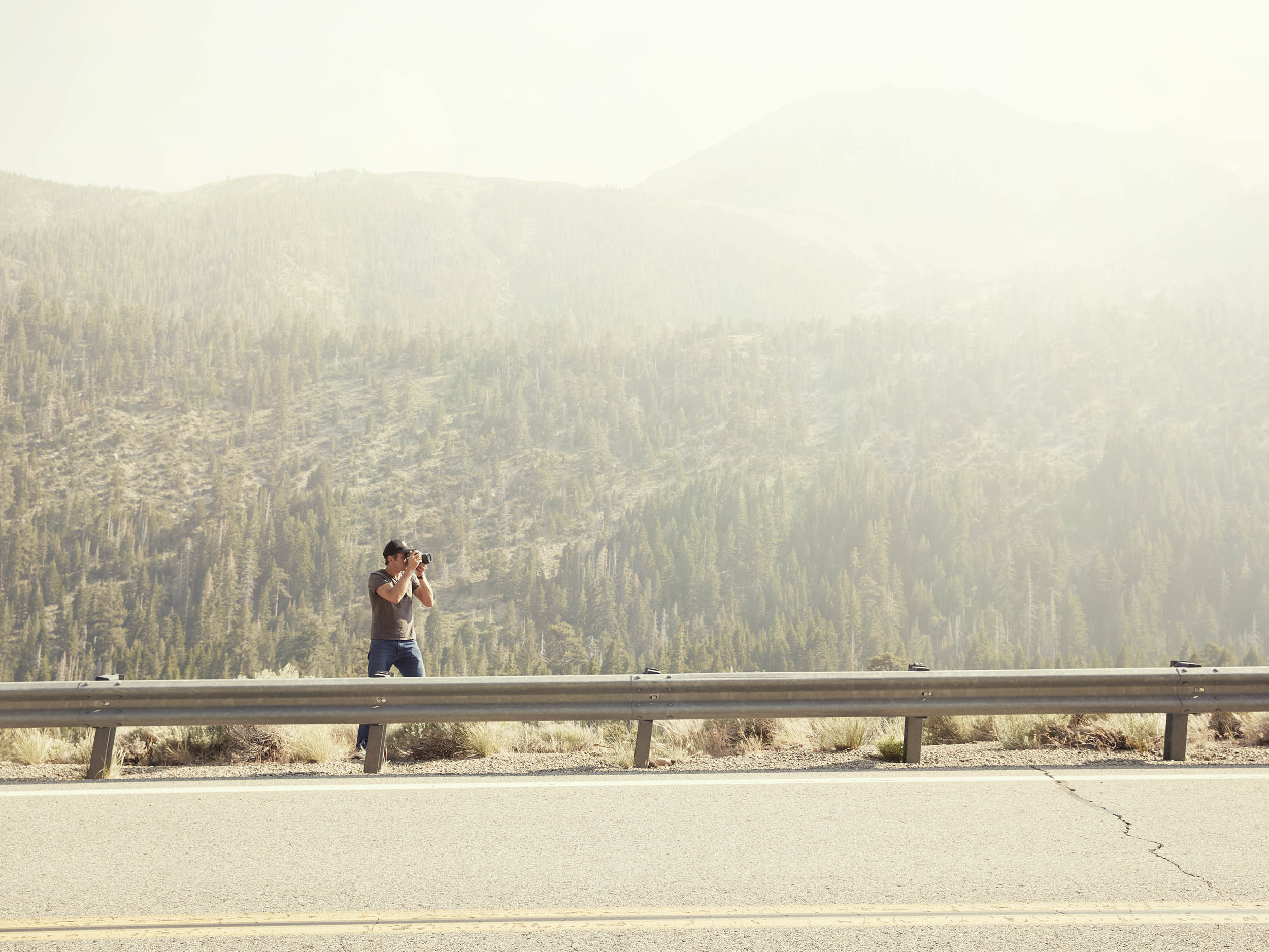 Person standing at roadside barrier photographing misty forested valley with golden morning light filtering through haze.