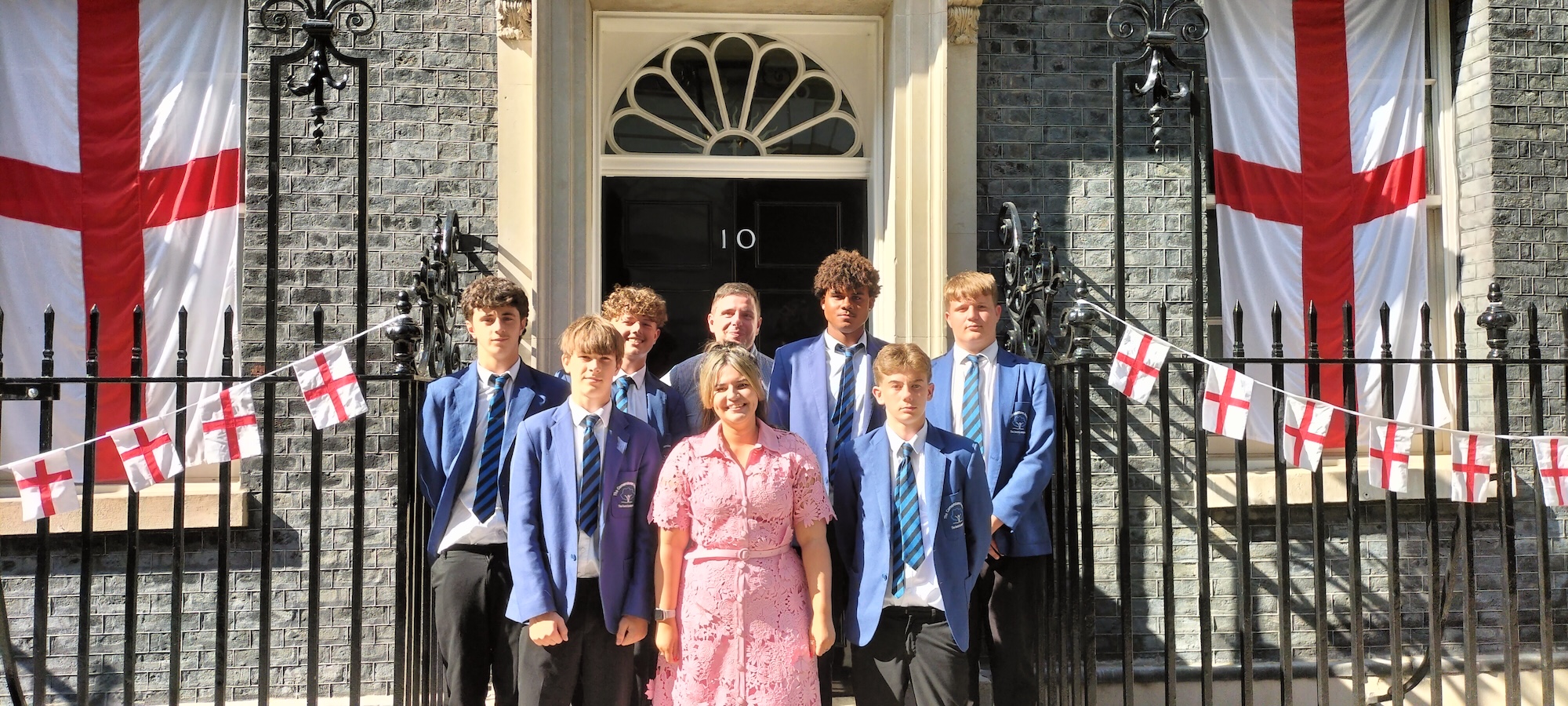 Six young people in uniform of Cornerstone Academy, Poole, stand with Dr A. Blower and F. Bartlett outside 10 Downing Street. Bunting and banners of the St George Cross flag hang either side of them.