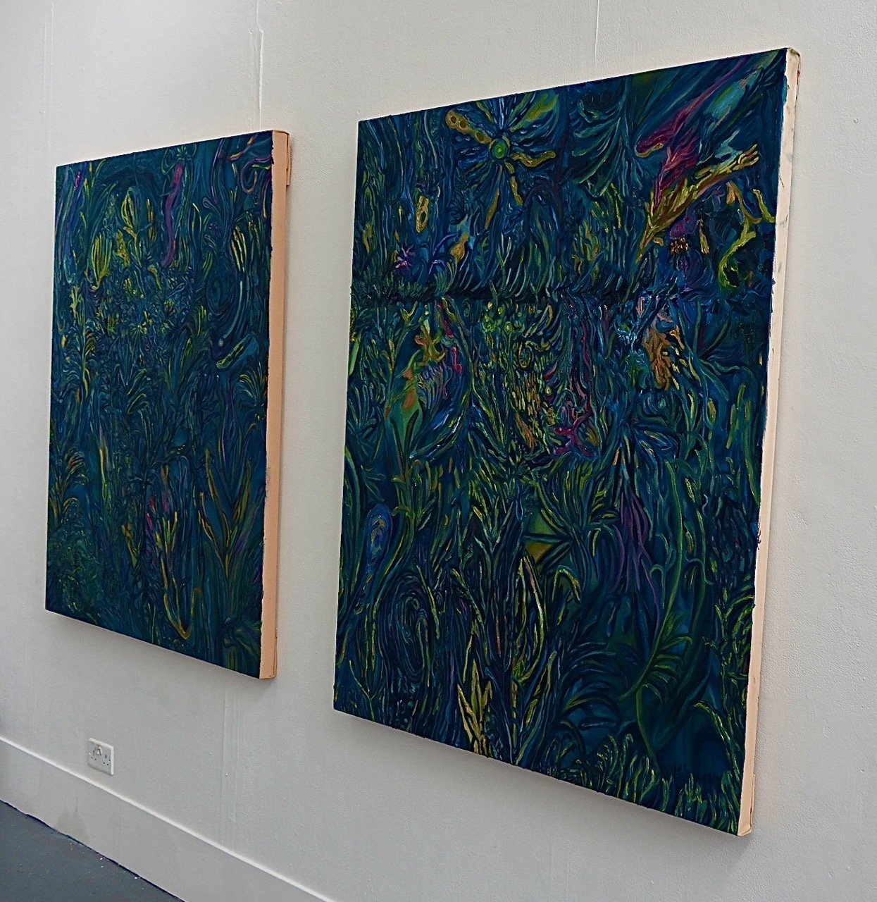 Two large abstract paintings with vibrant dark blue backgrounds and intricate, swirling patterns in various colours, displayed on a white wall. Each canvas is framed with a light wooden edge.