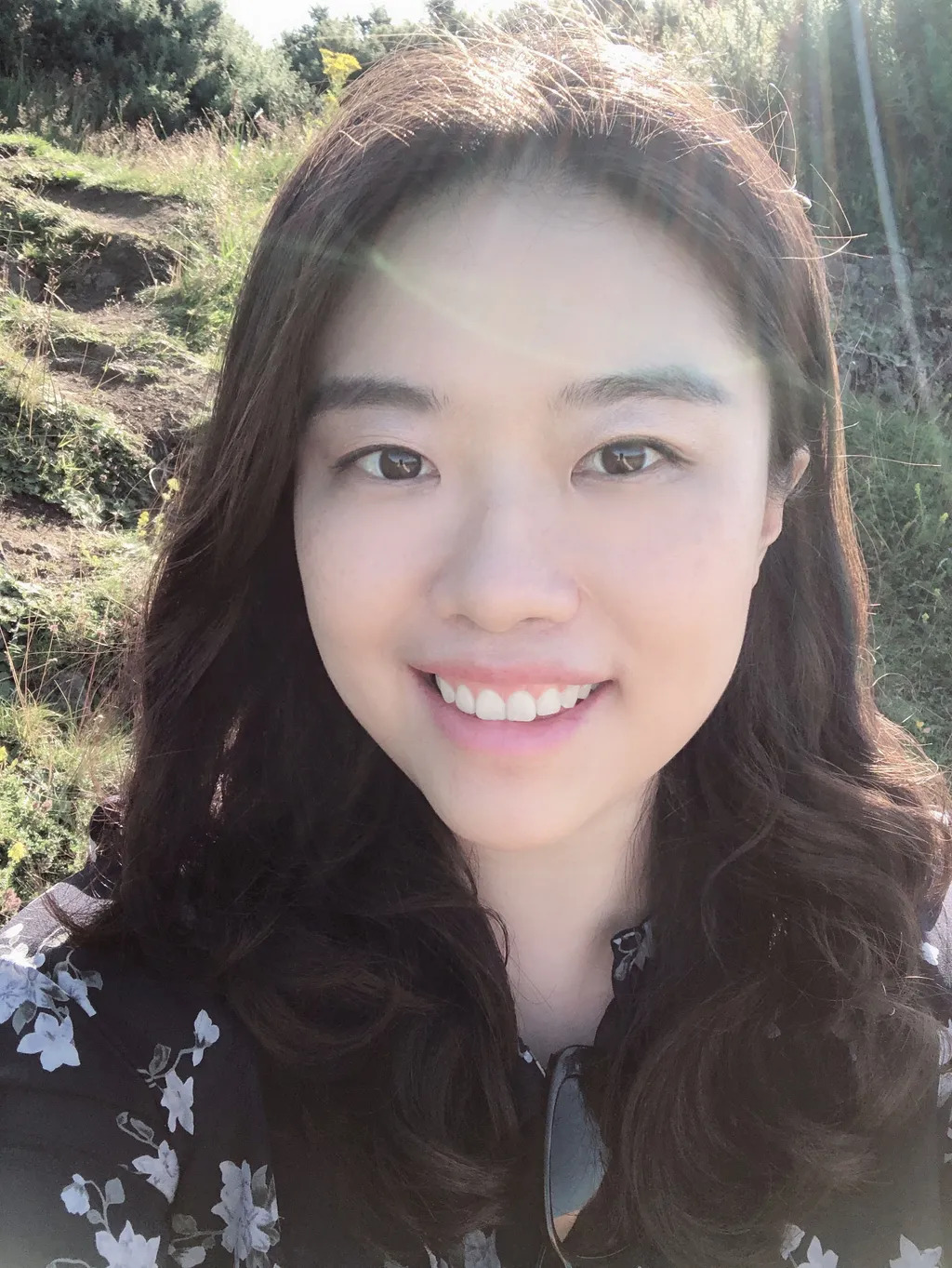 Smiling Asian woman with long dark hair wearing floral top, outdoors with rocky hillside and vegetation in background.