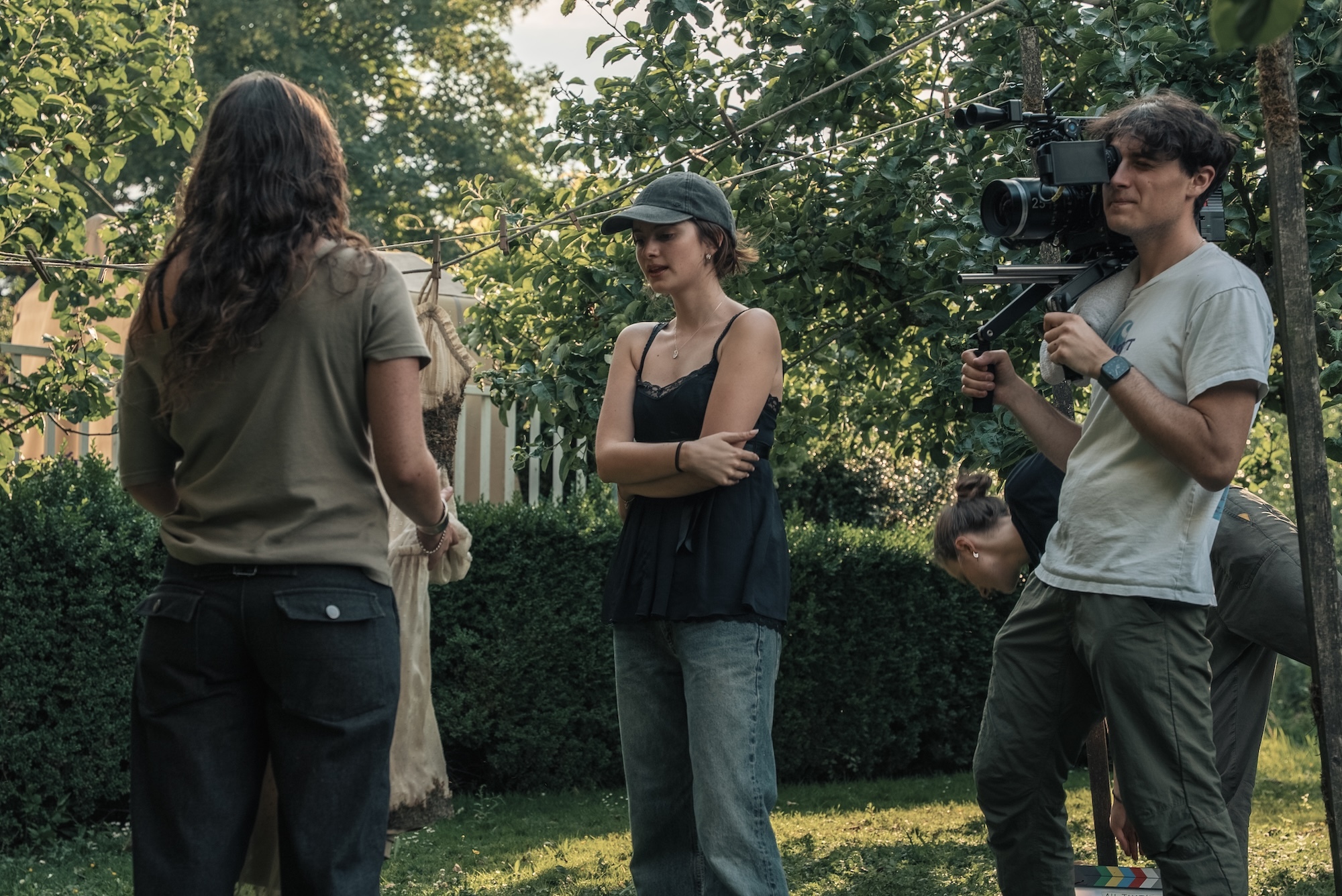 BA (Hons) Film Production graduate I. Campbell with two crew members and camera equipment recording two women in conversation outdoors amongst green trees and hedges in residential setting.