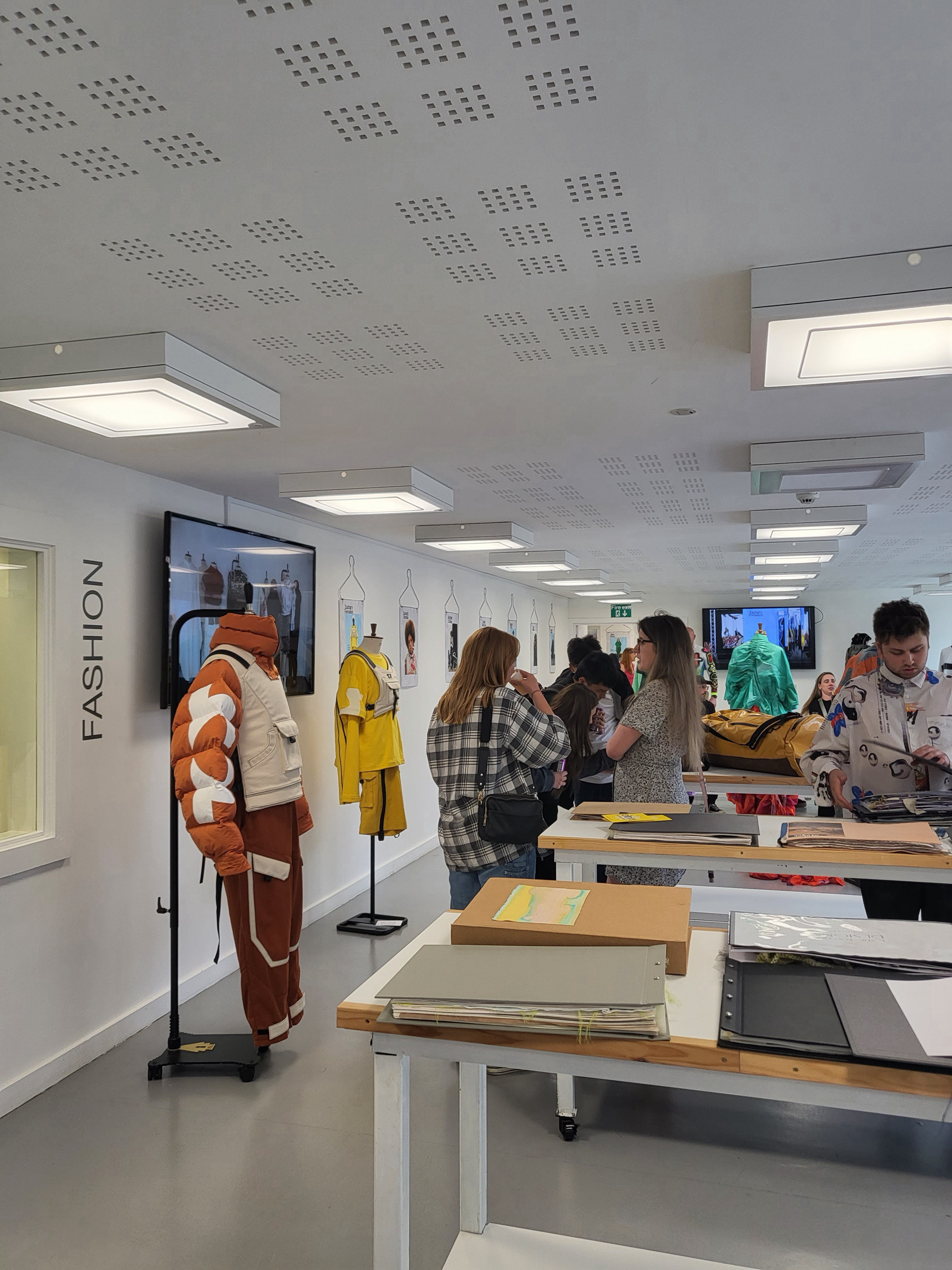 White classroom with perforated ceiling tiles, wooden desks, students working, fashion display with orange and yellow garments on stands