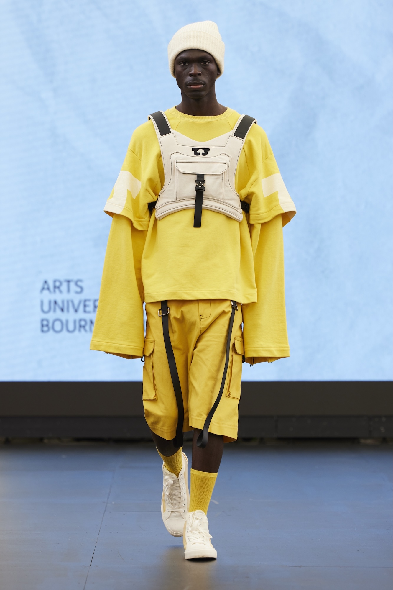 Model on runway wearing yellow layered outfit with white chest harness, white beanie and trainers against blue backdrop