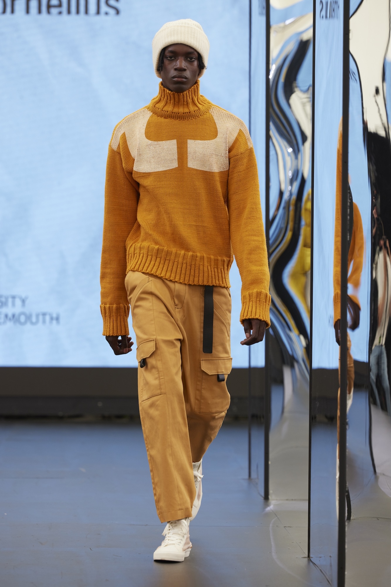 Man walking on runway wearing cream beanie, orange turtleneck jumper with cream panel detail, beige cargo trousers, white trainers