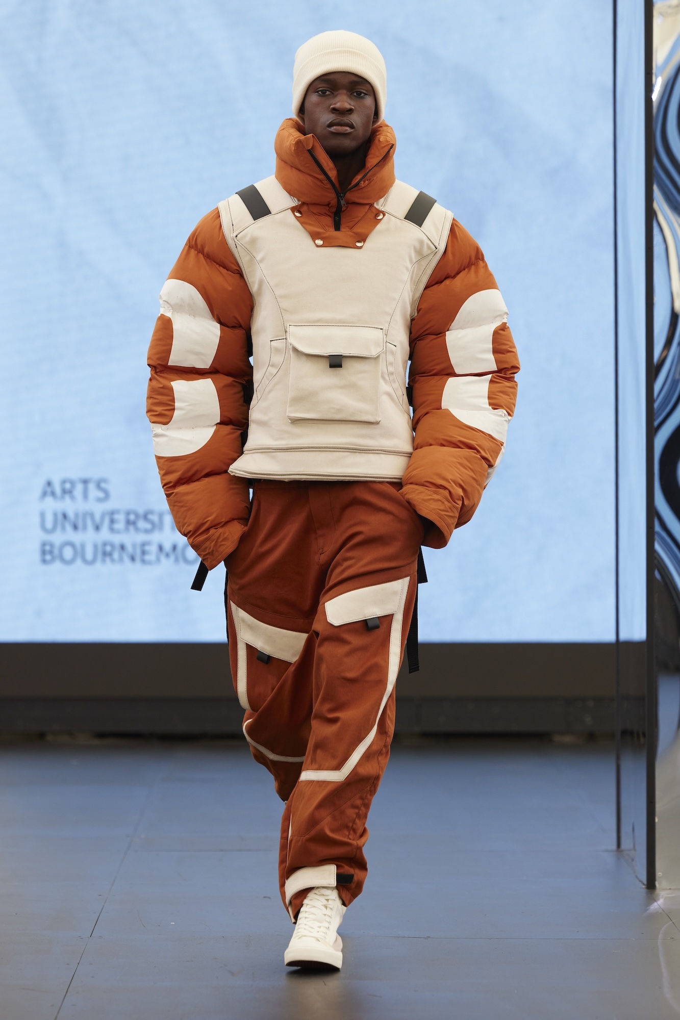 Male model on runway wearing white beanie, orange puffer jacket, cream vest with pockets, and orange trousers with white stripes