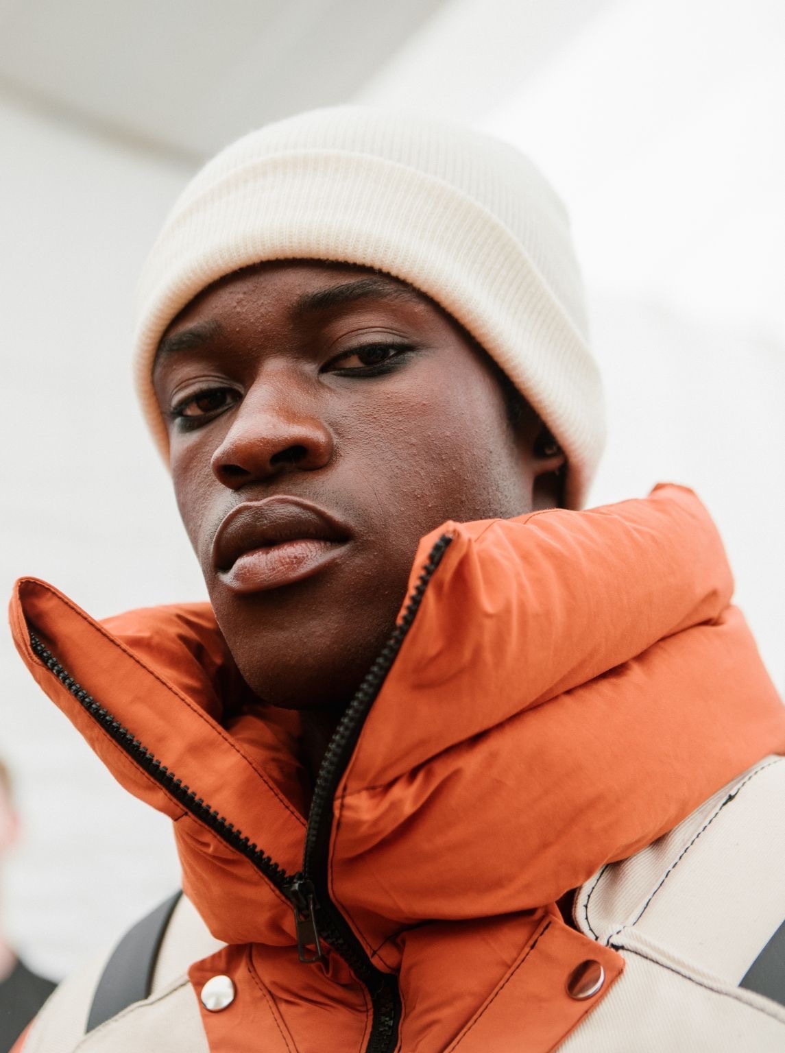 Black man wearing white knit beanie and bright orange puffer jacket, looking directly at camera against white background