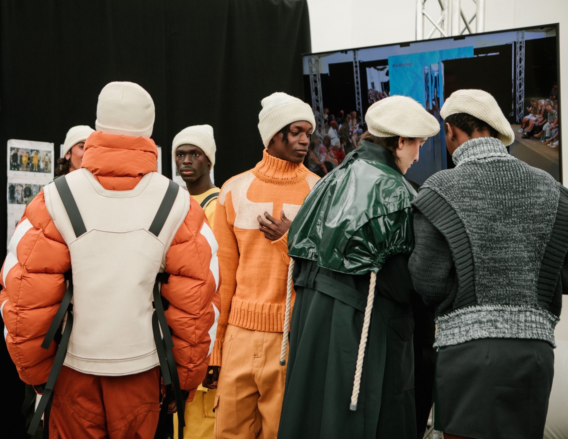 People wearing white caps viewing displays, dressed in orange and green clothing, standing in exhibition space with screens