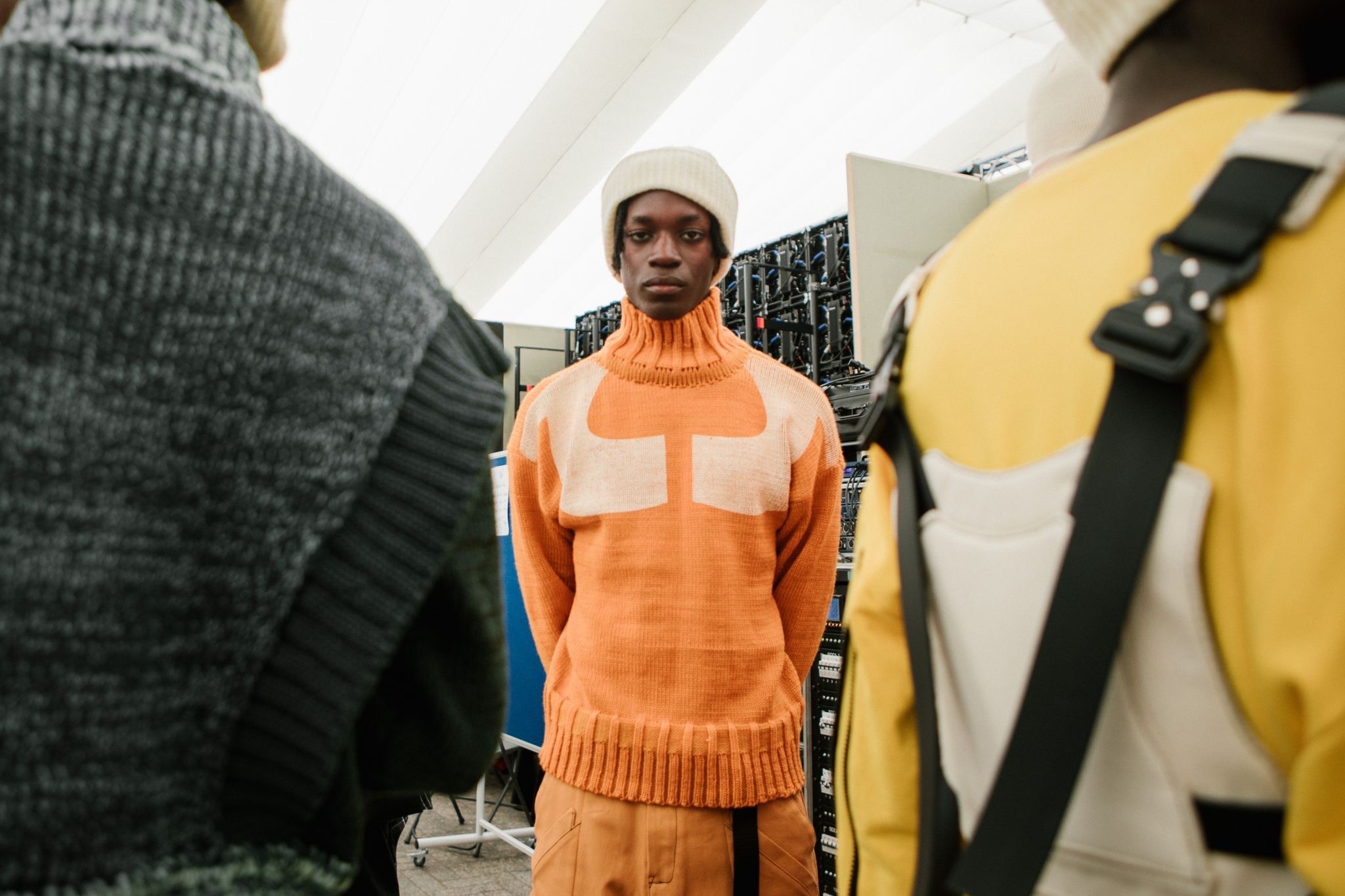 Person in orange knitted jumper with white cross design and white beanie stands between grey and yellow clothing displays