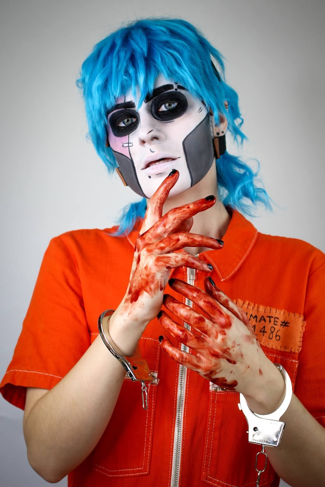 A person is dressed in a prison orange jumpsuit. They have blue hair and heavy eye make-up. There is blood on their hands and they're wearing a pair of broken handcuffs.