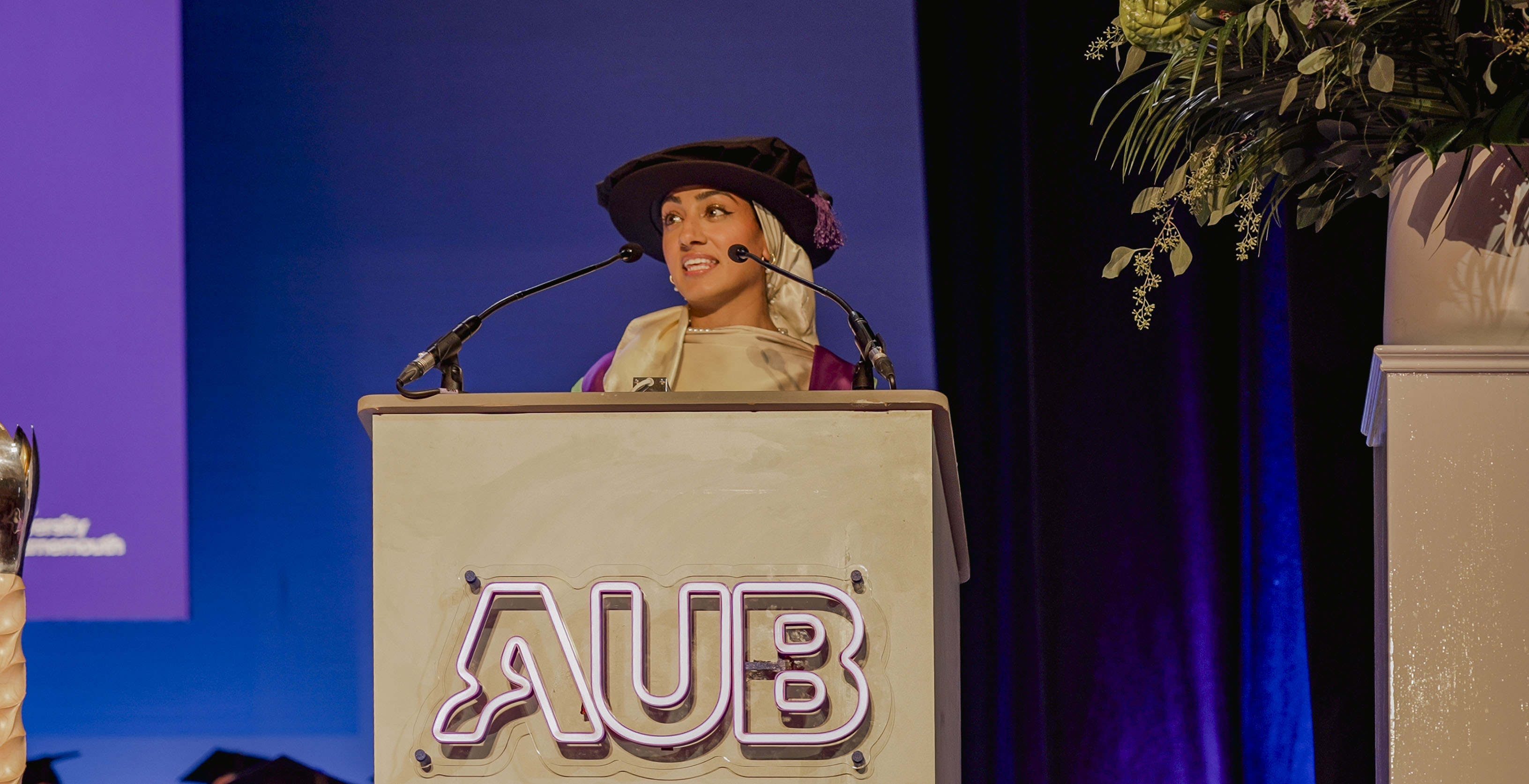 Z Jiwa speaks at a podium with an illuminated AUB sign, set against a blue-lit stage backdrop.