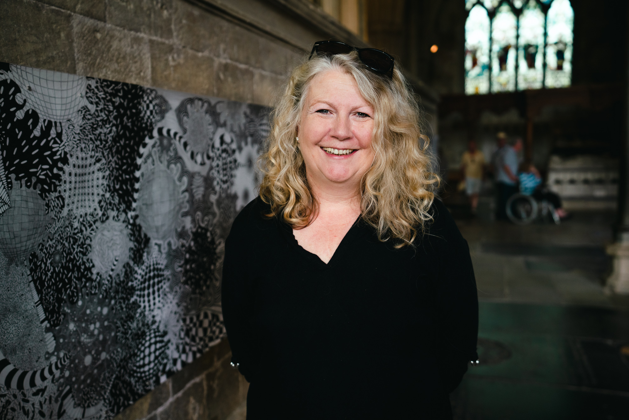 Artist Zara Saganic poses for a photo next to a long landscape canvas featuring black and white shapes, hung on a wall at Wells Cathedral.