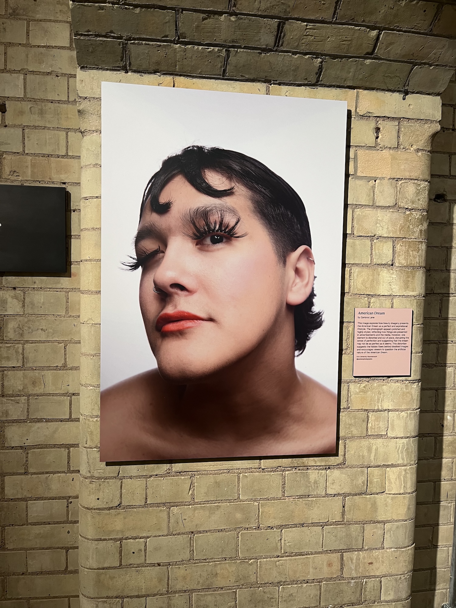 A large portrait photograph of a person with dramatic makeup and curled lashes mounted on a brick wall, with a small caption plaque beside it.