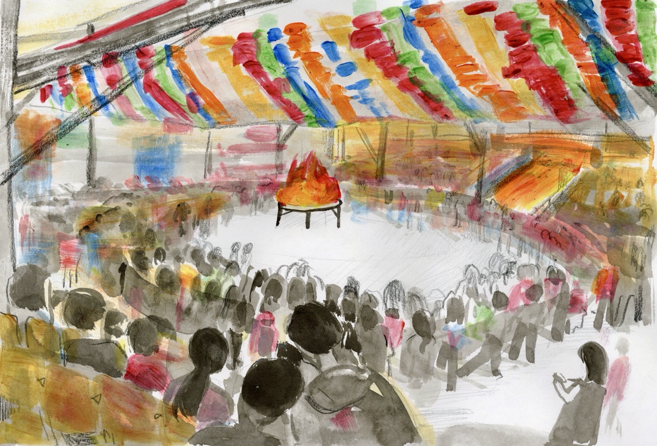 A crowd of people surrounds a central fire inside a large tent decorated with colourful hanging streamers and flags.