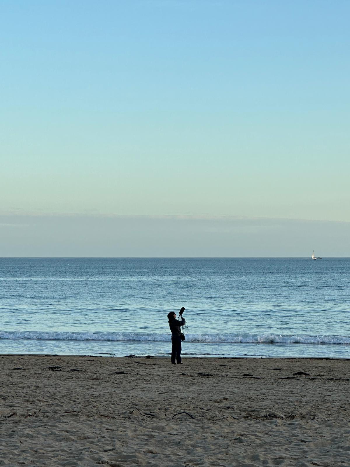 MA Film Practice student Z Chu standing on Bournemouth beach with the sea in the background, viewed from a distance. They are holding up a boom microphone.