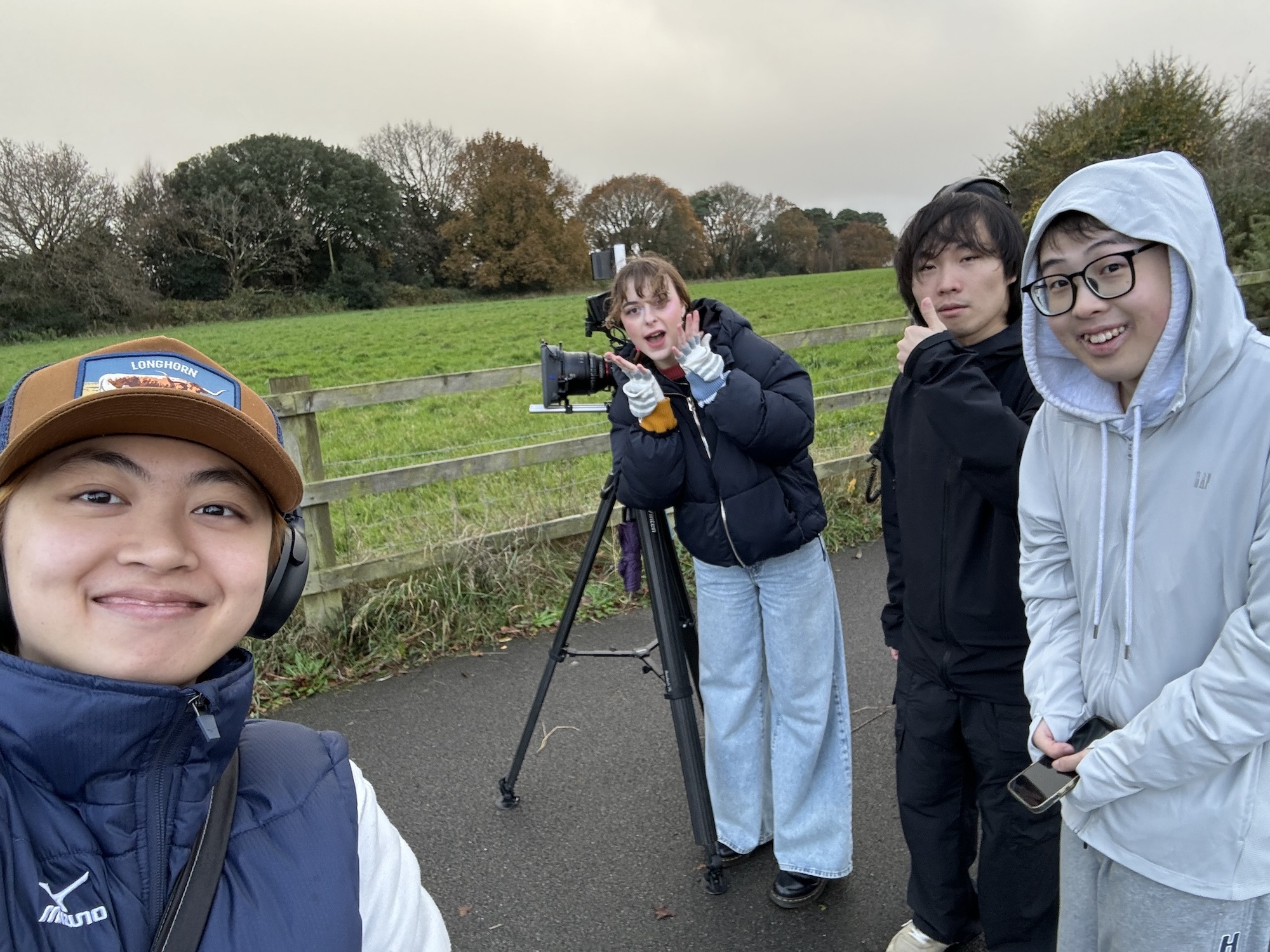 Four MA Film Practice students pose for a photo outside in the rain. A field and a camera on a tripod appear in the background.