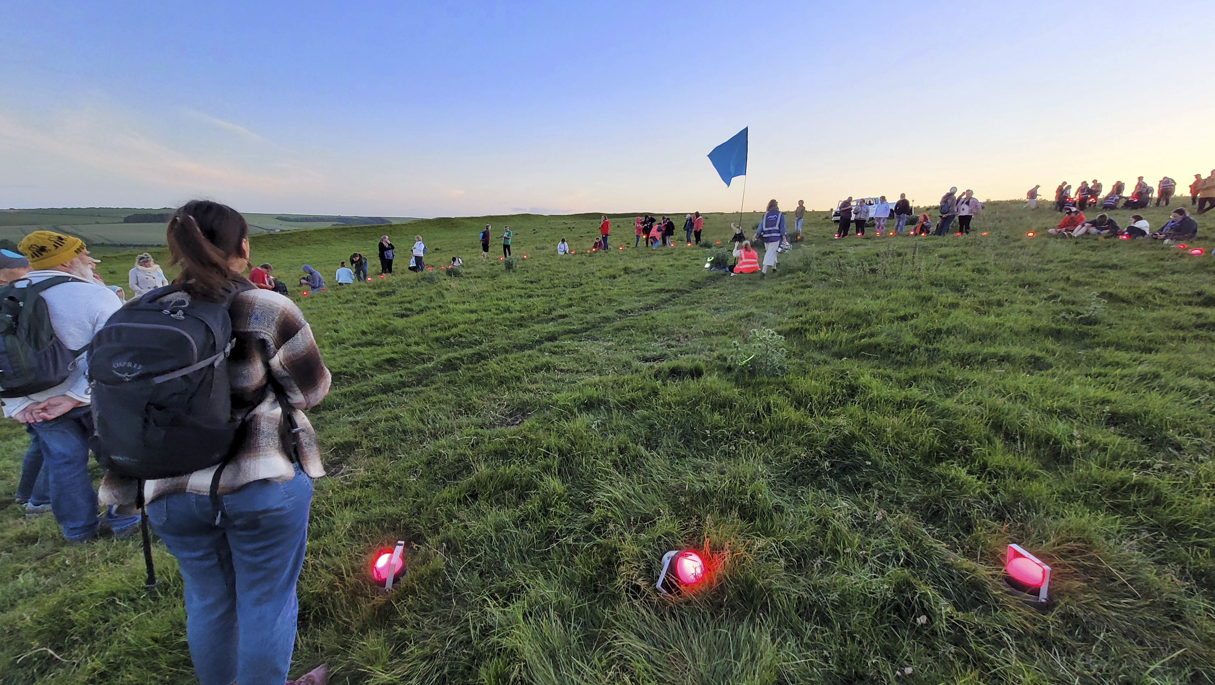 A large group of people gathers in a green field with red lanterns in front of them